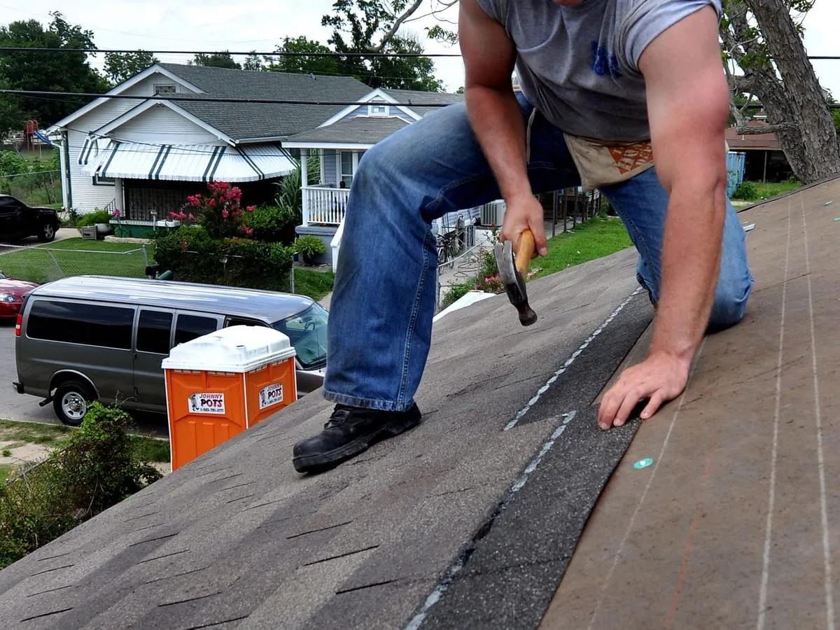 Volunteer kneeling on a roof, nailing asphalt shingles over underlayment with a hammer