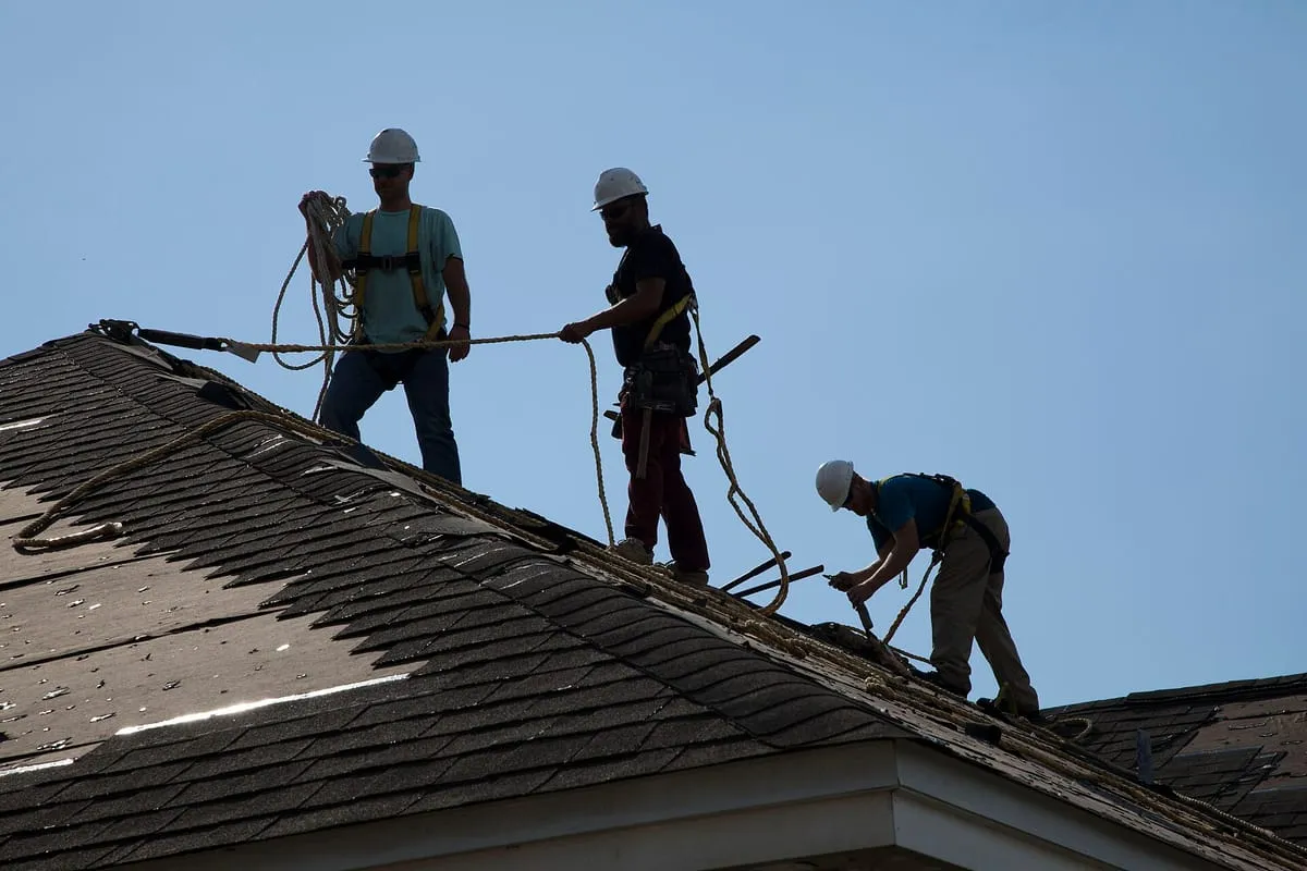 Three workers in hard hats and safety harnesses on a damaged shingle roof