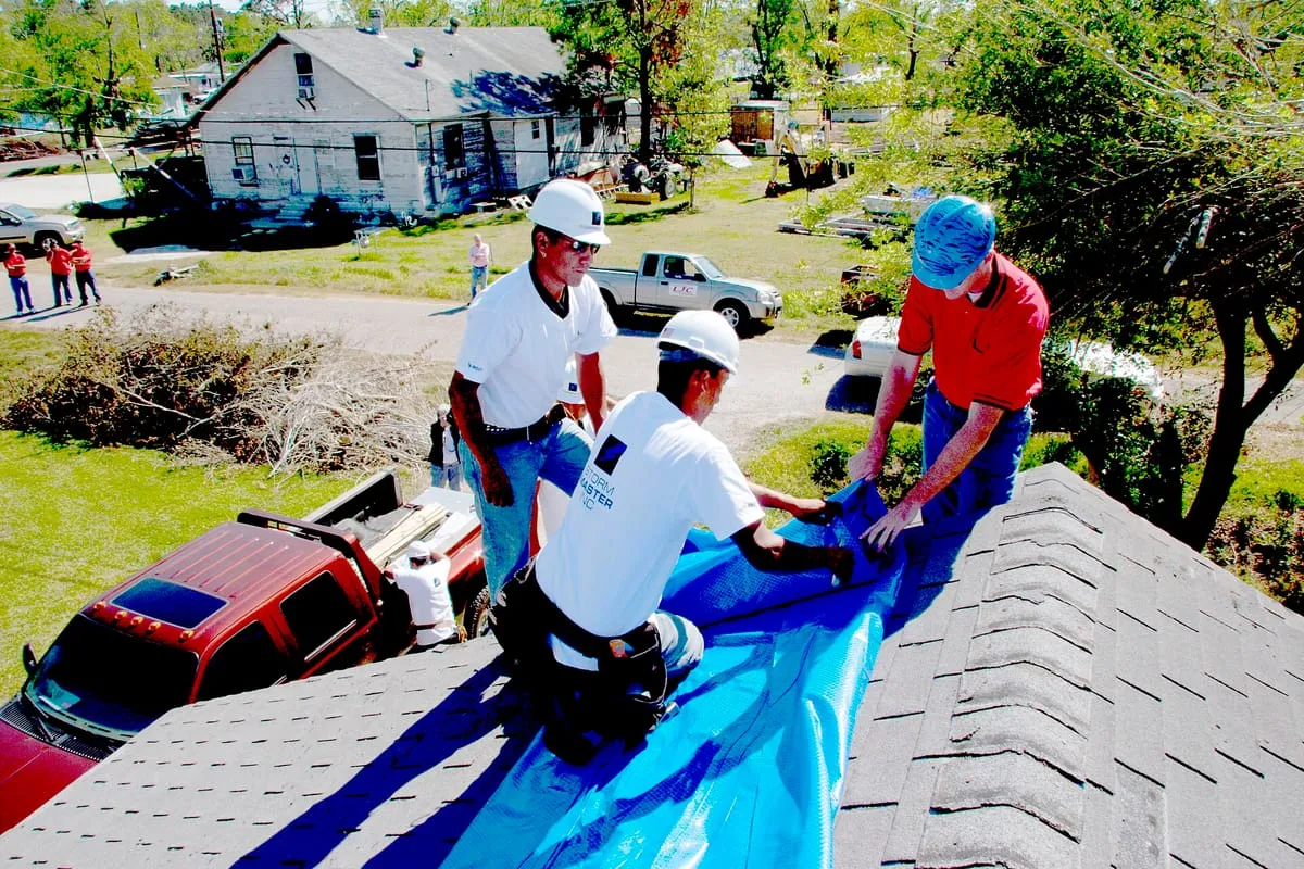 Three workers in hard hats spreading blue sheeting over a damaged shingle roof