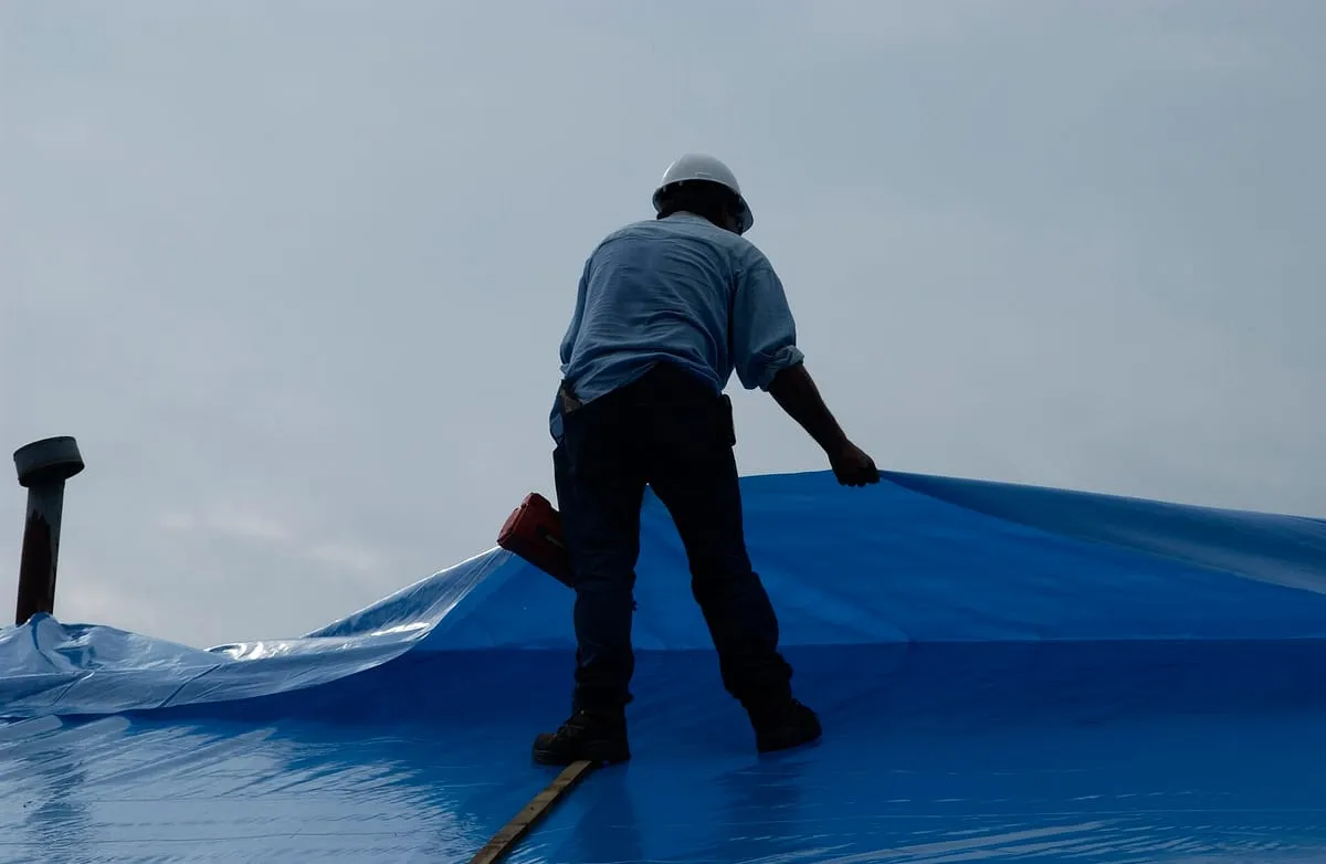 Worker in a hard hat pulling blue plastic sheeting across a roof under gray skies