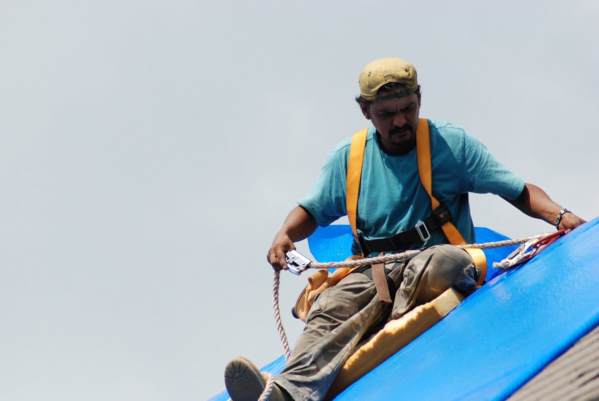 Worker in a harness sitting on a roof covered with blue tarp, holding a safety line