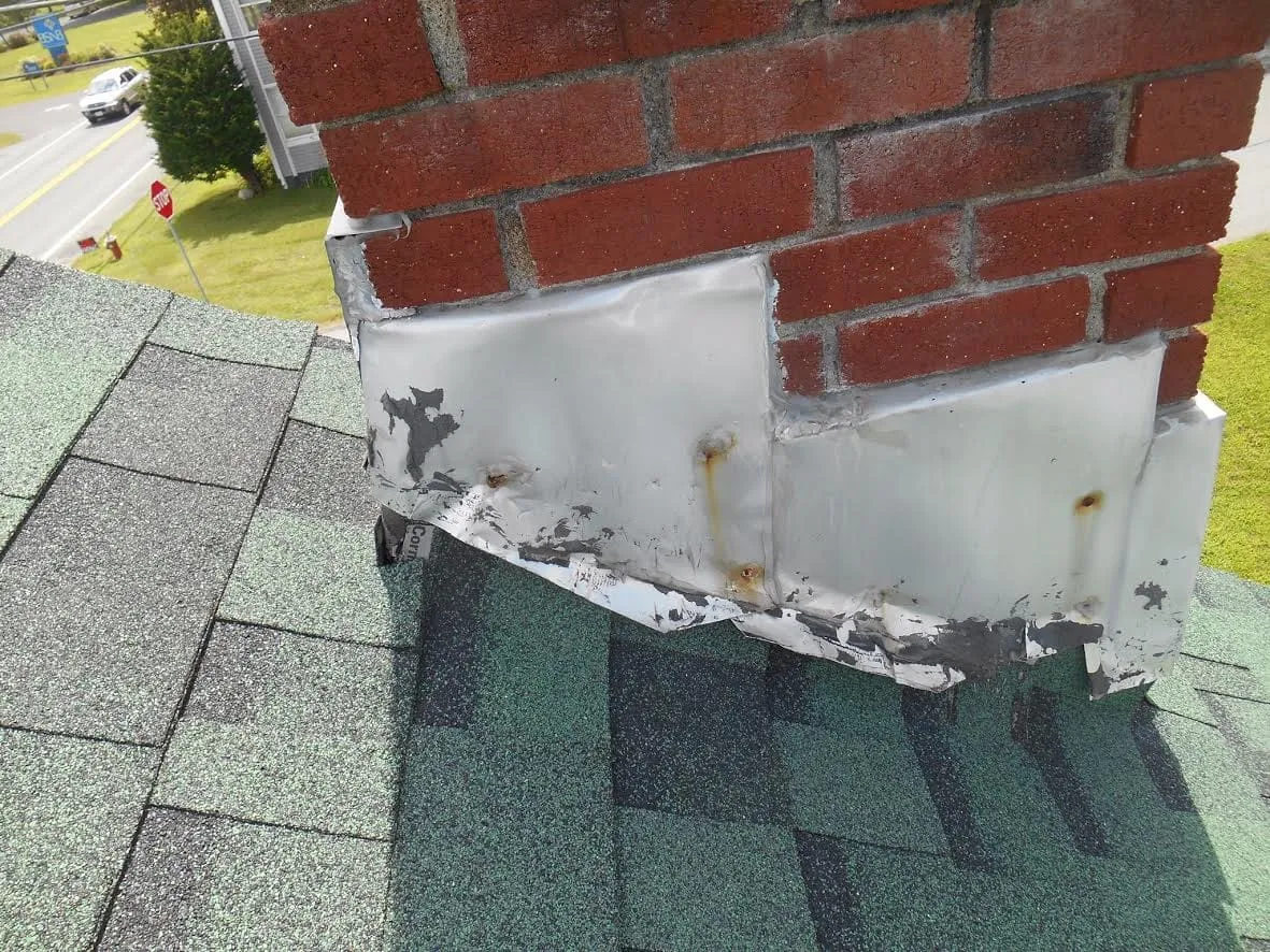 Rusted, poorly fitted metal flashing around a brick chimney on a shingle roof