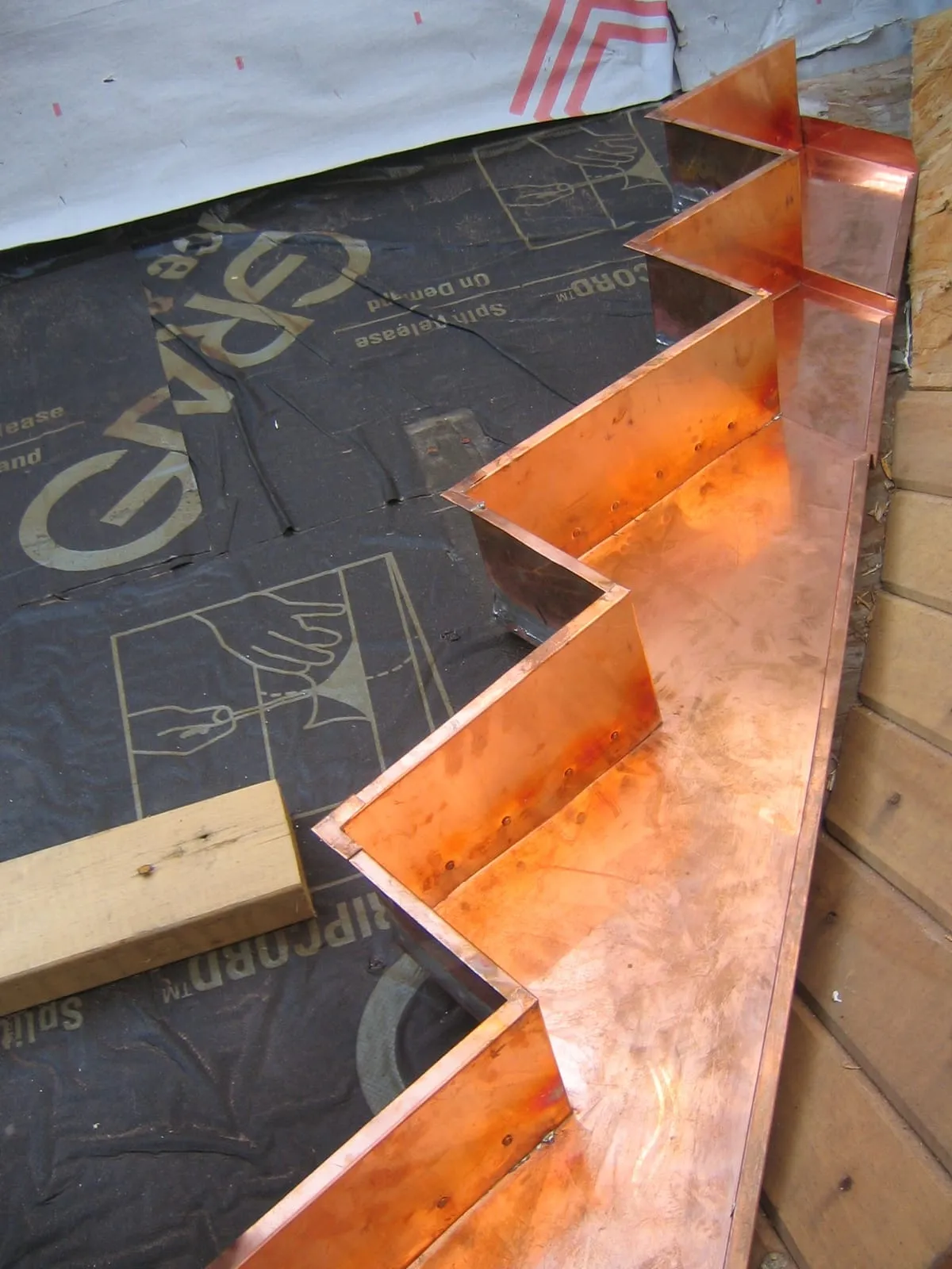 Copper stepped flashing fitted against a brick chimney over black roof underlayment
