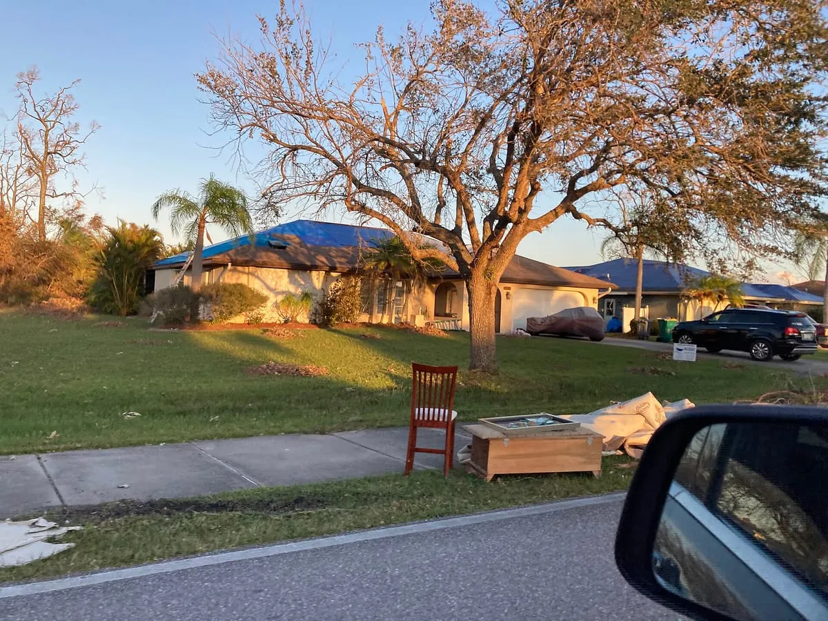 Single-story house with a blue tarp on its roof and damaged furniture at the curb