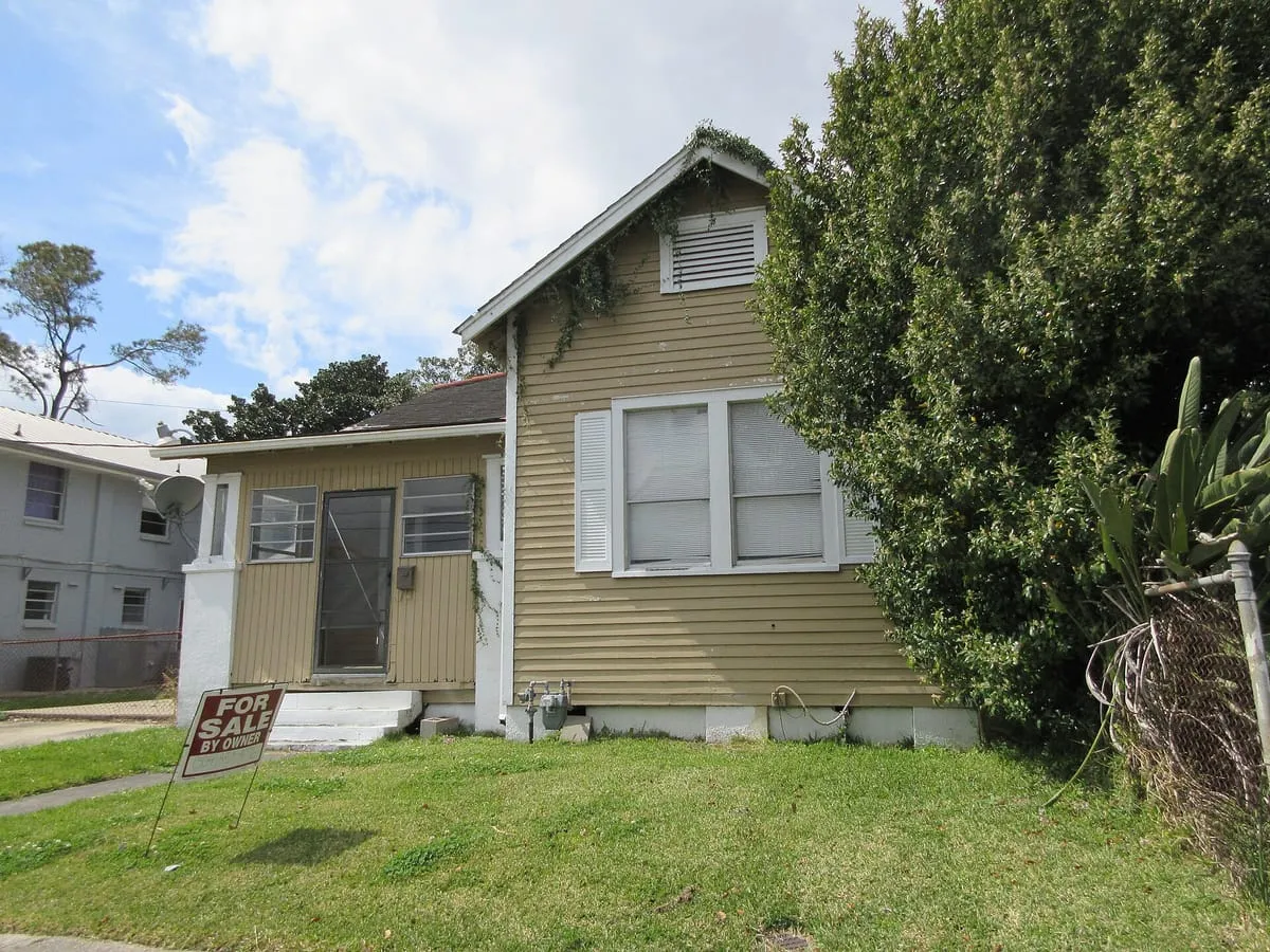 Small one-story house with siding and a for sale by owner sign in the yard