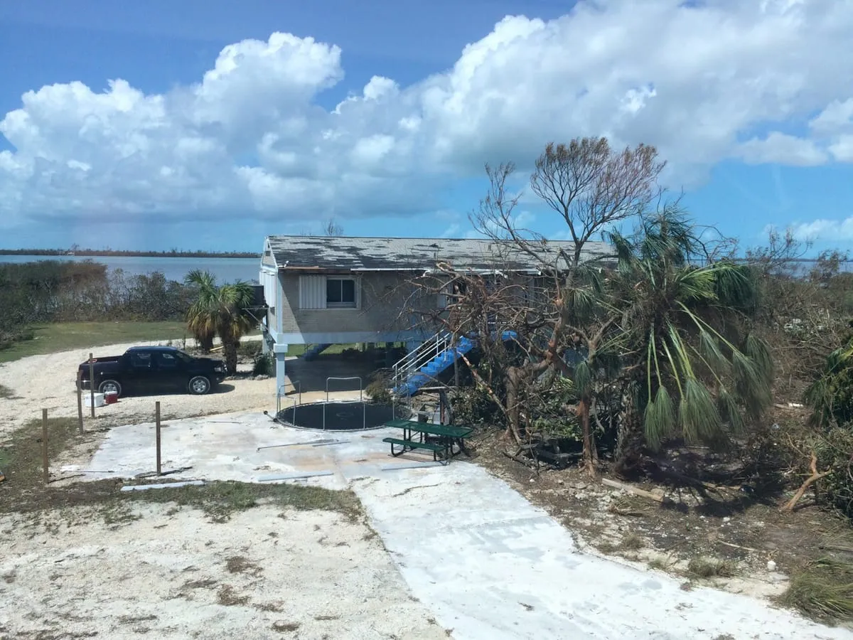 Raised building with patches of missing shingles, palm trees and water behind it
