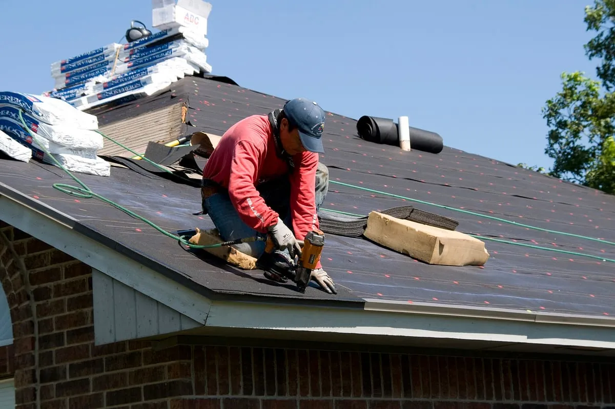 Roofer kneeling on underlayment nailing shingles, with bundles stacked near the ridge