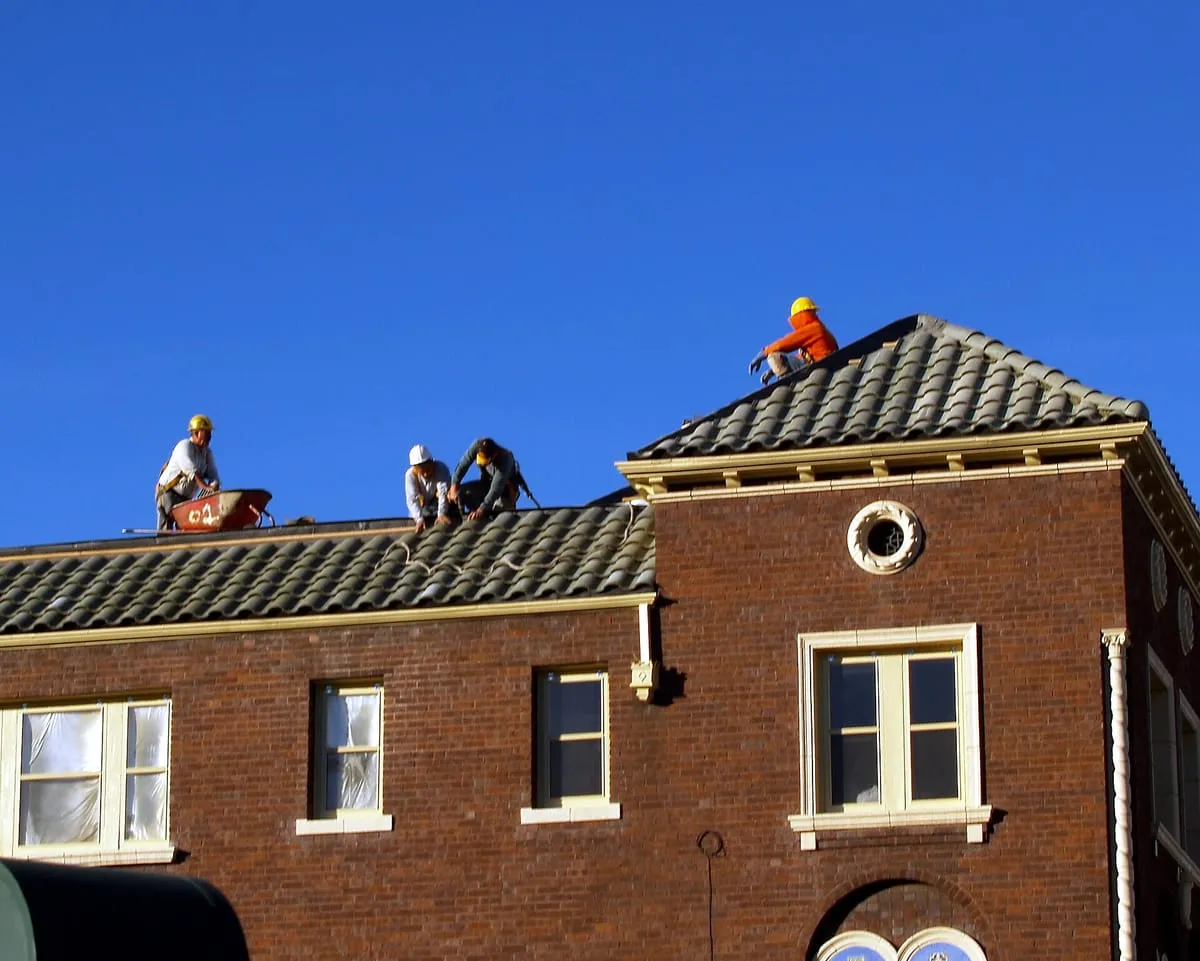 Roofers in hard hats working on the tile roof of a brick building