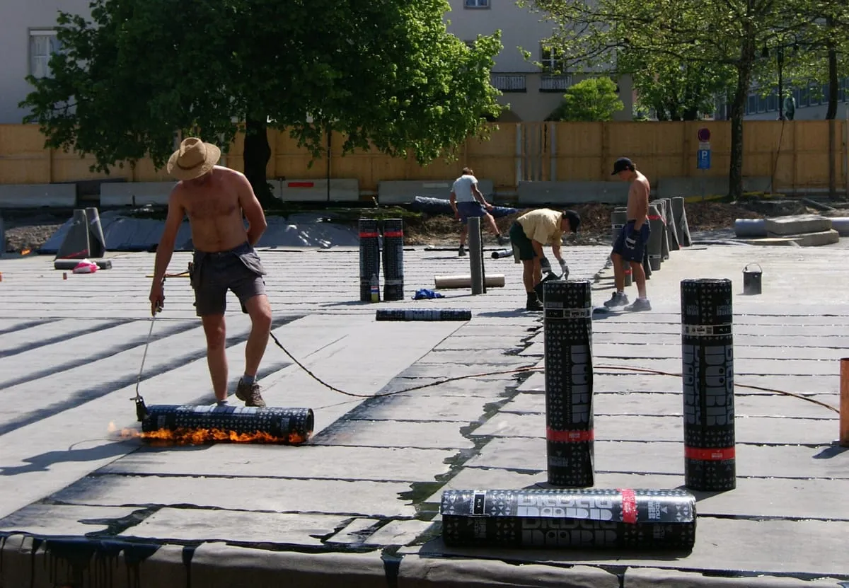 Workers torch-applying rolls of bitumen roofing membrane across a large flat roof