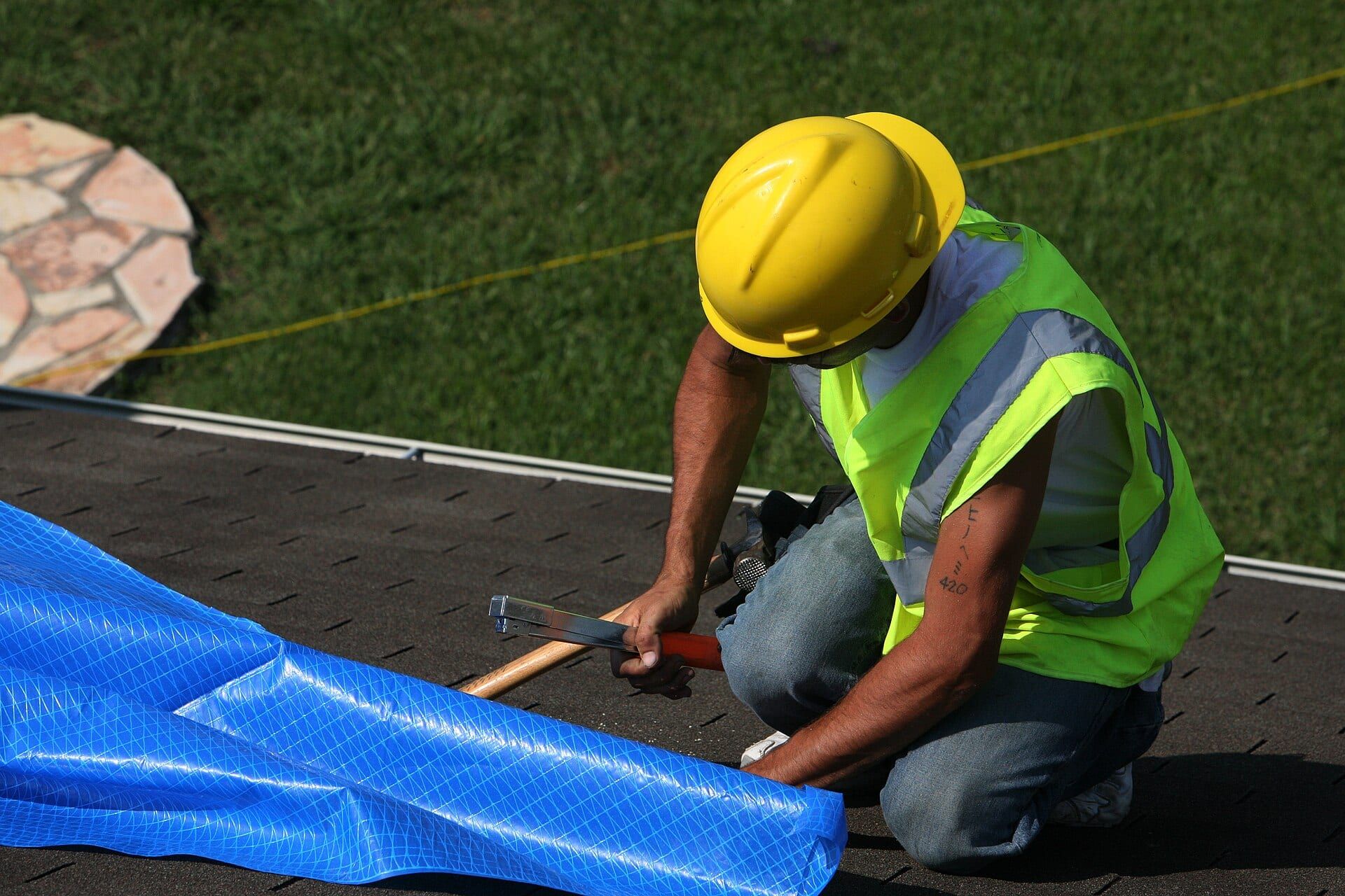 Worker in a hard hat and safety vest nailing down a blue tarp on a shingle roof